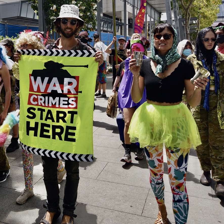 People in colourful costumes holding a banner saying "War Crimes Start Here"